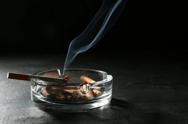 在灰色桌子上的玻璃烟灰缸中 在黑色背景下燃烧着香烟 案文的篇幅 — 图库照片