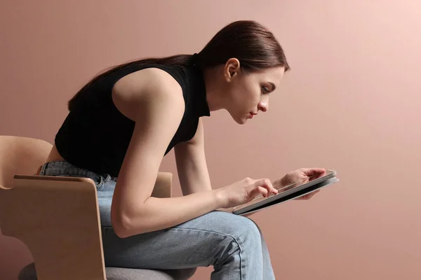 Young woman with bad posture using tablet while sitting on chair against pale pink background