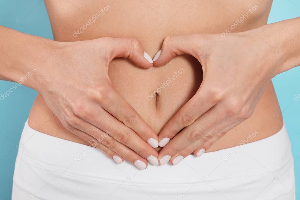 Woman making heart with hands on her belly against light blue background, closeup