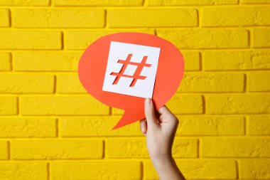 Sarı tuğla duvara hashtag sembollü kırmızı kağıt konuşma balonu tutan kadın, yakın plan.