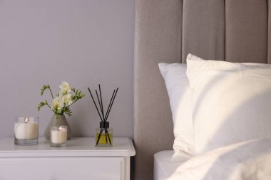 Aromatik kamış oda spreyi, frezya çiçekleri ve yatak odasındaki beyaz komodindeki mumlar.