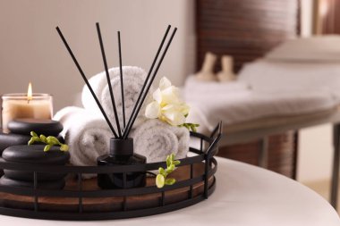 Aromatik kamış oda spreyi, rulo havlular, spa taşları ve beyaz masa üzerinde mum.