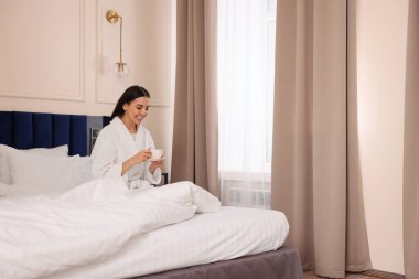 Otel odasında bornoz giymiş, yatakta kahve içen mutlu genç bir kadın.