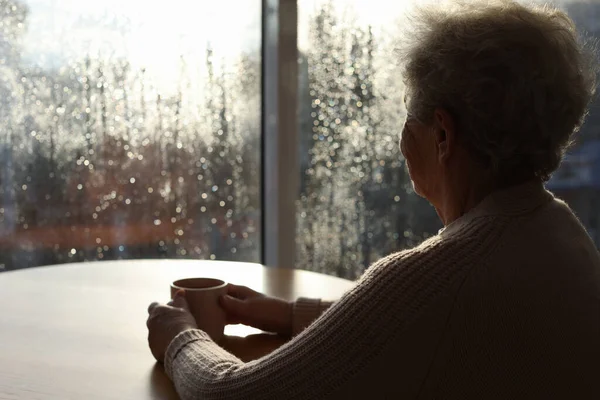 Starsza Kobieta Kubkiem Napoju Pobliżu Okna Deszczowy Dzień Miejsce Tekst — Zdjęcie stockowe