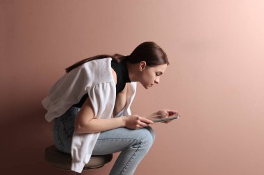Tablet kullanan, soluk pembe arka planda taburede oturan kötü duruşu olan genç bir kadın.