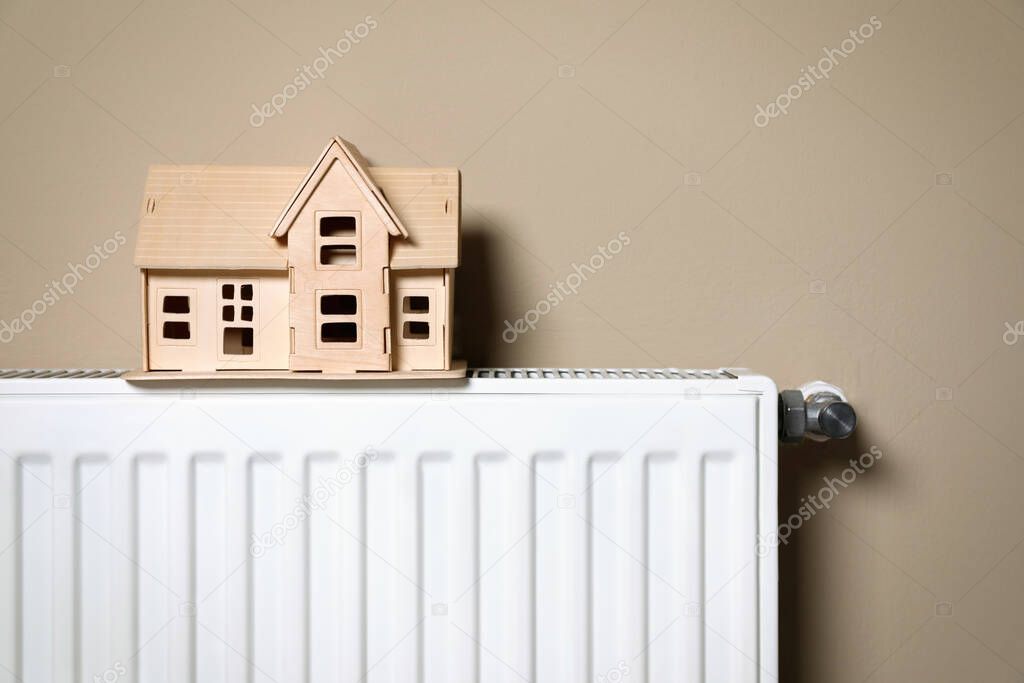 Modern radiator with wooden house near beige wall indoors. Winter heating efficiency