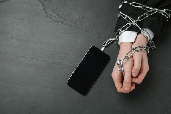 Человек, показывающий прикованные руки к смартфону за черным столом, вид сверху. Интернет-зависимость