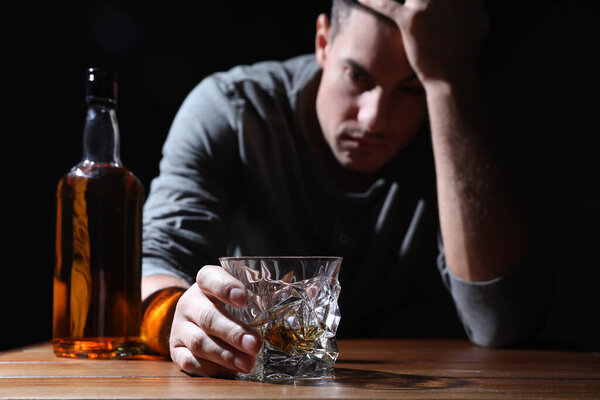 Зависимый человек за деревянным столом на черном фоне, сосредоточьтесь на стакане алкогольного напитка