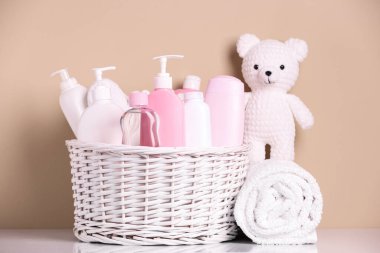 Bebek kozmetik ürünleri, banyo aksesuarları ve bej arka planda beyaz masa üzerine örülmüş oyuncak ayı sepetleri.