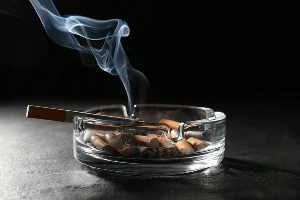 在黑色背景下灰色桌子上的烟灰缸里燃烧着香烟 — 图库照片
