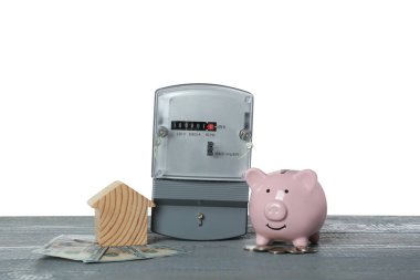 Elektrik sayacı, ev modeli, para ve kumbara gri ahşap masada beyaz arka planda.