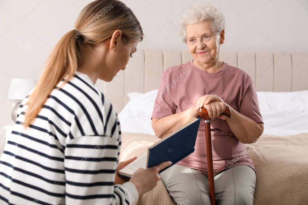 Young caregiver reading book to senior woman in bedroom. Home care service
