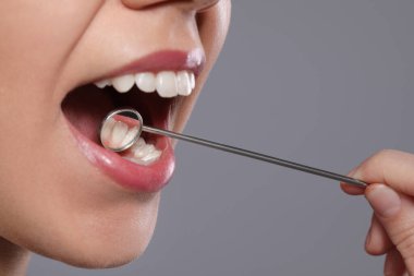 Gri arka planda ayna olan dişçi kadının dişlerini inceliyor.