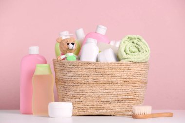 Bebek kozmetik ürünleri, banyo aksesuarları ve hasır sepetteki oyuncak pembe arka planda beyaz masada