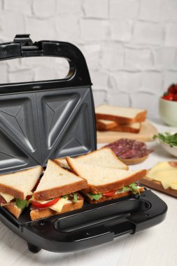 Lezzetli sandviçler ve beyaz ahşap masa malzemeleriyle modern ızgara makinesi.