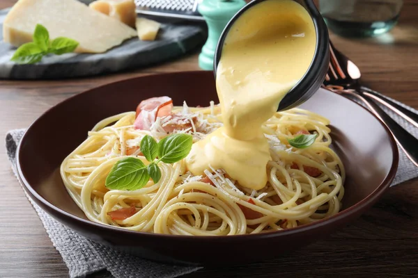 Pouring tasty cheese sauce onto spaghetti with meat on wooden table, closeup