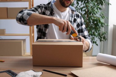 İçerideki ahşap masada paket açmak için bıçak kullanan adam, yakın plan.