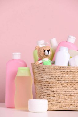 Bebek kozmetik ürünleri, banyo aksesuarları ve hasır sepetteki oyuncak pembe arka planda beyaz masada