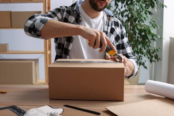 İçerideki ahşap masada paket açmak için bıçak kullanan adam, yakın plan.