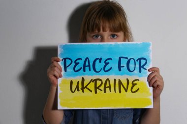 Beyaz duvarın yanında Ukrayna için barış yazılı kağıt tutan üzgün küçük kız