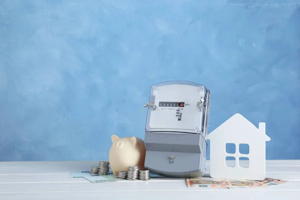 Electricity meter, piggy bank, house model and money on white wooden table. Space for text