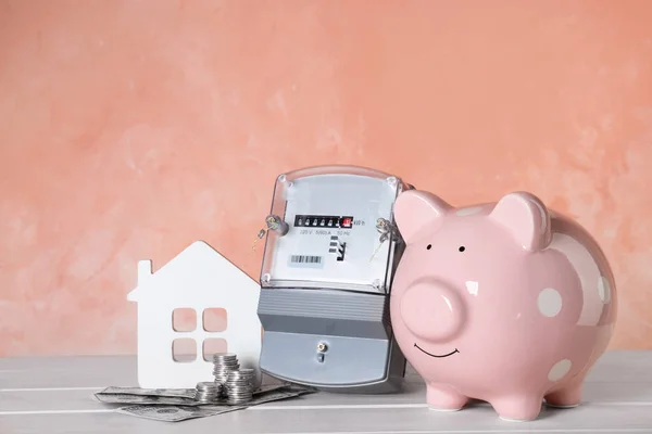 Electricity meter, piggy bank, house model and money on white wooden table