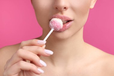 Dudakları şekerle kaplı genç bir kadın pembe arka planda lolipop yiyor, yakın plan.