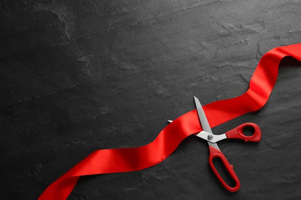 Red ribbon and scissors on black background, top view. Space for text