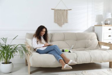 Not defteri ve tabletle evdeki kanepede ders çalışan Afro-Amerikalı kadın. Uzaklık öğrenimi