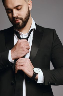 Yakışıklı sakallı adam gri arka planda kravatını sıkıyor.
