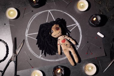 Gri masa üzerinde iğne ve tören malzemeleriyle delik deşik edilmiş kadın voodoo bebeği.