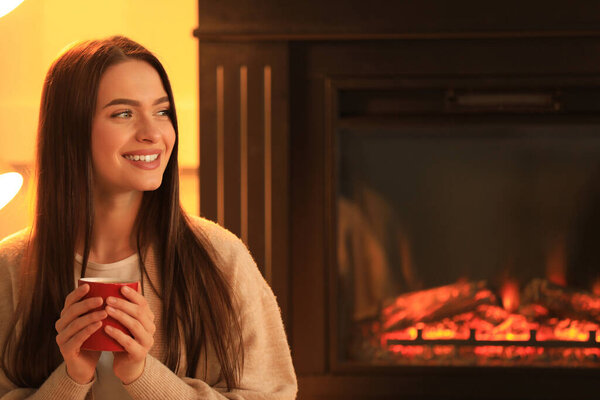 Young woman with cup of hot drink near fireplace indoors. Cozy atmosphere