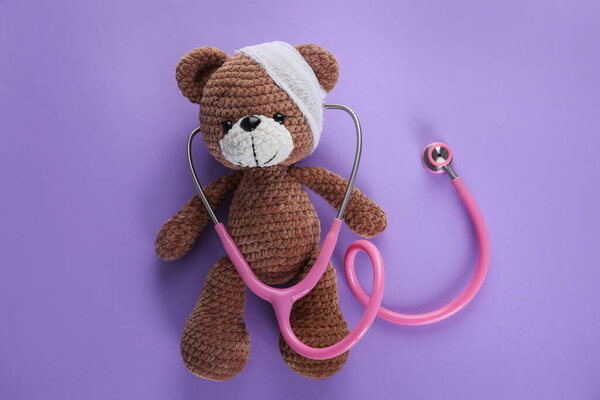 Cute toy bear with bandage and stethoscope on violet background, top view. Children's hospital
