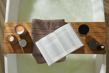 Ahşap banyo tepsisi, açık kitap, bir bardak şarap ve kozmetik ürünler küvette, üst manzara