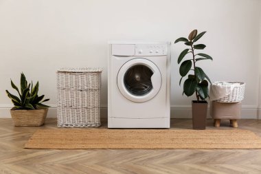 Çamaşır odasının içinde modern çamaşır makinesi ve beyaz duvarın yanında hasır sepet var.