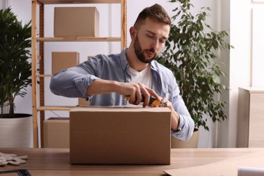 İçerideki ahşap masada paket açmak için bıçak kullanan bir adam.