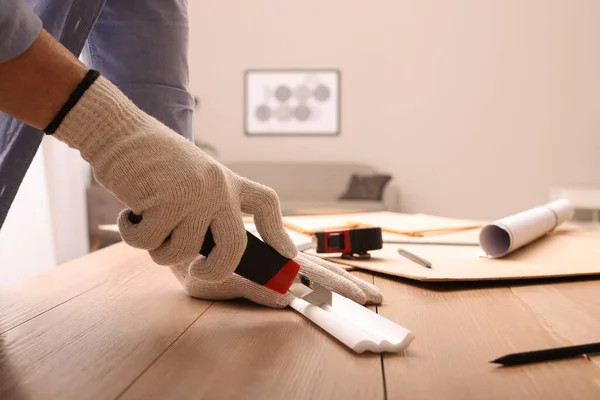 Man cutting foam crown molding with utility knife at wooden table indoors, closeup