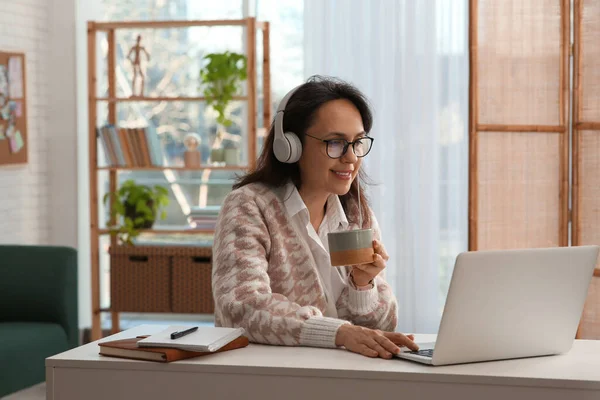 現代のラップトップとヘッドフォンを持つ女性は テーブルで屋内で学びながらお茶を飲む — ストック写真