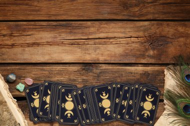 Tarot kartları, tavus kuşu tüyleri, değerli taşlar ve ahşap masa üzerindeki eski kitap, yazı için yeri olan üst manzara. Ters taraf