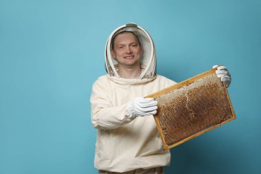 Üniformalı arı yetiştiricisi açık mavi arka planda bal peteğiyle kovan çerçevesi tutuyor.