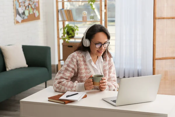 現代のラップトップとヘッドフォンを持つ女性は テーブルで屋内で学びながらお茶を飲む — ストック写真