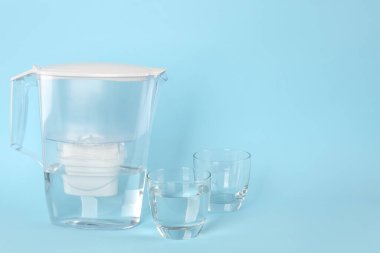 Açık mavi arka planda arıtılmış suyla filtre sürahisi ve bardaklar. Metin için boşluk