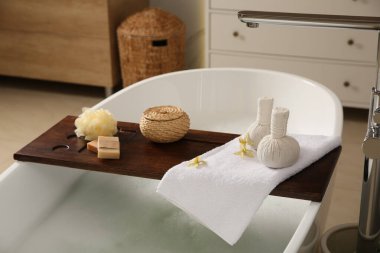 Ahşap banyo tepsisi, bitkisel masaj torbaları ve banyo konforları küvette.
