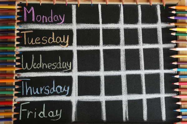 Colorful pencils and drawn weekly school timetable on black chalkboard, top view