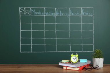 Çalar saat, kırtasiye ve yeşil karatahtanın yanındaki ahşap masa üzerinde çizilmiş okul zaman çizelgesi.
