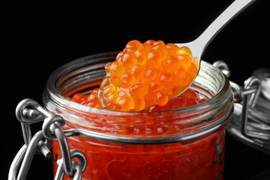 Cam kavanoz ve kaşık, siyah arka planda lezzetli kırmızı havyar, yakın plan.