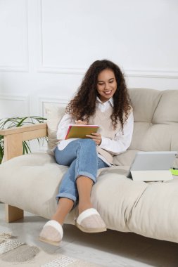 Not defteri ve tabletle evdeki kanepede ders çalışan Afro-Amerikalı kadın. Uzaklık öğrenimi