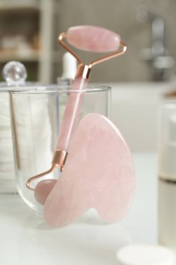 Gül kuvars gua sha aracı, doğal yüz silindiri ve beyaz masa üzerindeki tuvalet malzemeleri.