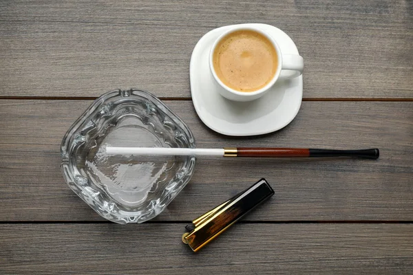 Ashtray with long cigarettes holder, lighter and cup of coffee on wooden table, flat lay