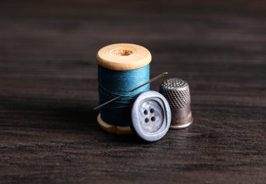 Spool of light blue thread with needle, button and thimble on wooden table. Sewing accessories
